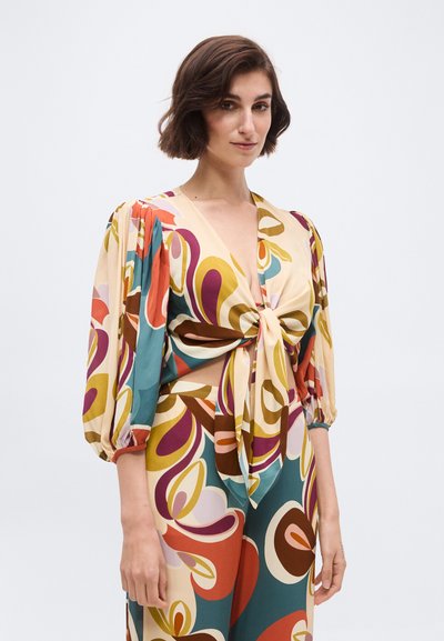 Γυναίκα με κοντά καστανά μαλλιά που φοράει πολύχρωμο μπλουζάκι με ρετρό μοτίβο, φουσκωτά μανίκια και δεμένο κόμπο μπροστά.