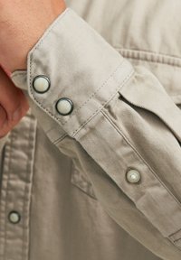 Beige langwerpige shirtmanchet met twee knoopsluitingen, een in licht plastic en een in donker metaal, met zichtbare stikdetails.