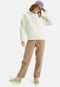 Crème-kleurige fleece hoodie met een kangoeroezak, verstelbaar koord en blauwe logo. Gecombineerd met lichtbruine corduroy broek en beige schoenen.