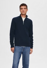 Marinblå zip-up tröja med hög krage, texturerad tyg, ribbade ärmslut och nederkant, bärs över en ljusfärgad skjorta och är kombinerad med jeans.