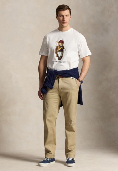 Mann trägt weißes T-Shirt mit Bär-Grafik, beige Hose, marineblauen Pullover um die Taille gebunden und blaue Turnschuhe, steht vor neutralem Hintergrund.