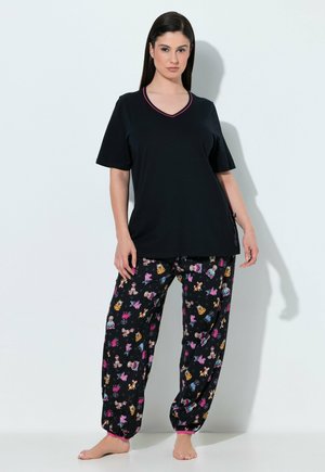 Woman wearing black short-sleeve top and loose black pants with colorful cartoon character prints, standing barefoot against a white wall.