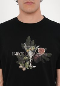 Czarny t-shirt z graficznym wzorem przedstawiającym kieliszek koktajlowy, oliwki i figi, z zielonymi liśćmi oraz napisem "EMPORIO ARMANI" w kolorze srebrnym.