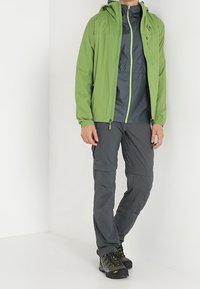 Vihreä vedenpitävä takki hupulla, kontrastina harmaa paita alla, harmaat cargo-housut ja mustat vaelluskengät, joissa on vihreitä yksityiskohtia.