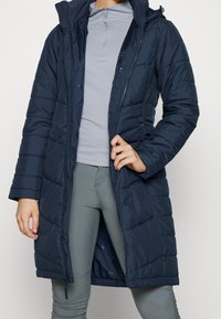 Tmavě modrý péřový kabát s kapucí, zapínáním na zip vpředu, bočními kapsami a délkou po kolena. Nositelný přes šedé tričko s dlouhým rukávem a legíny.