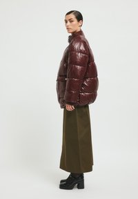 Glanzende bordeaux pufferjas met een hoge kraag, met een boxy ontwerp en gestikte panelen. Gecombineerd met een lange olijfgroene rok en dikke zwarte schoenen.