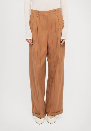 Pantalons à jambes larges en tissu marron chaud avec des rayures verticales. Équipés de plis et d'un ourlet retroussé. Associés à des chaussures pointues blanches cassées.