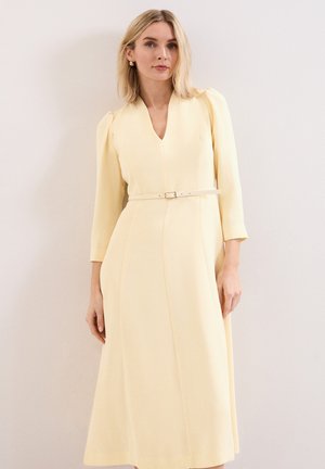 Femme blonde portant une longue robe jaune pâle avec un col en V, des manches trois-quarts et une fine ceinture à la taille, debout contre un mur uni.