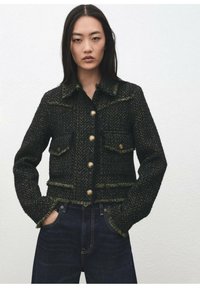 Czarna i zielona kurtka w jodełkę z postrzępionymi krawędziami, kołnierzem klapowym i dużymi złotymi guzikami, wyposażona w dwie przednie kieszenie.
