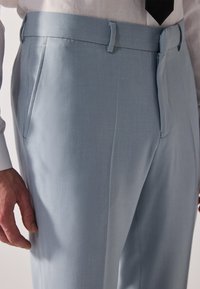 Los pantalones de traje azul claro presentan una textura suave, corte recto, trabillas para el cinturón y bolsillos laterales, combinados con una camisa blanca y una corbata negra.