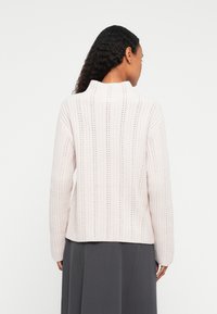 Žena s dlouhými tmavými kudrnatými vlasy, oblečená ve světle růžovém pleteném svetru a tmavě šedé sukni, zobrazená zezadu na bílém pozadí.