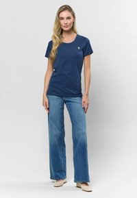 Sötétkék póló kerek nyakkal és rövid ujjakkal, bal mellen egy kis hímzett logóval. Világoskék harangszárú farmerral párosítva.