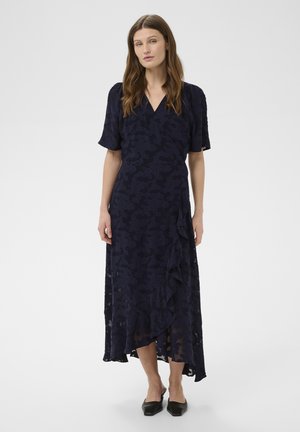 Naine, kes kannab tumel sinist lillelist mähkimiskleiti lühikeste varrukatega ja volangidega, seljas mustad terava ninaga kingad, seisab valge tausta ees.