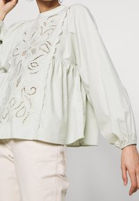 Hellgrüne Baumwollbluse mit Puffärmeln, Rüschenausschnitt und aufwändig gesticktem Ausschnittmuster auf der Vorderseite.