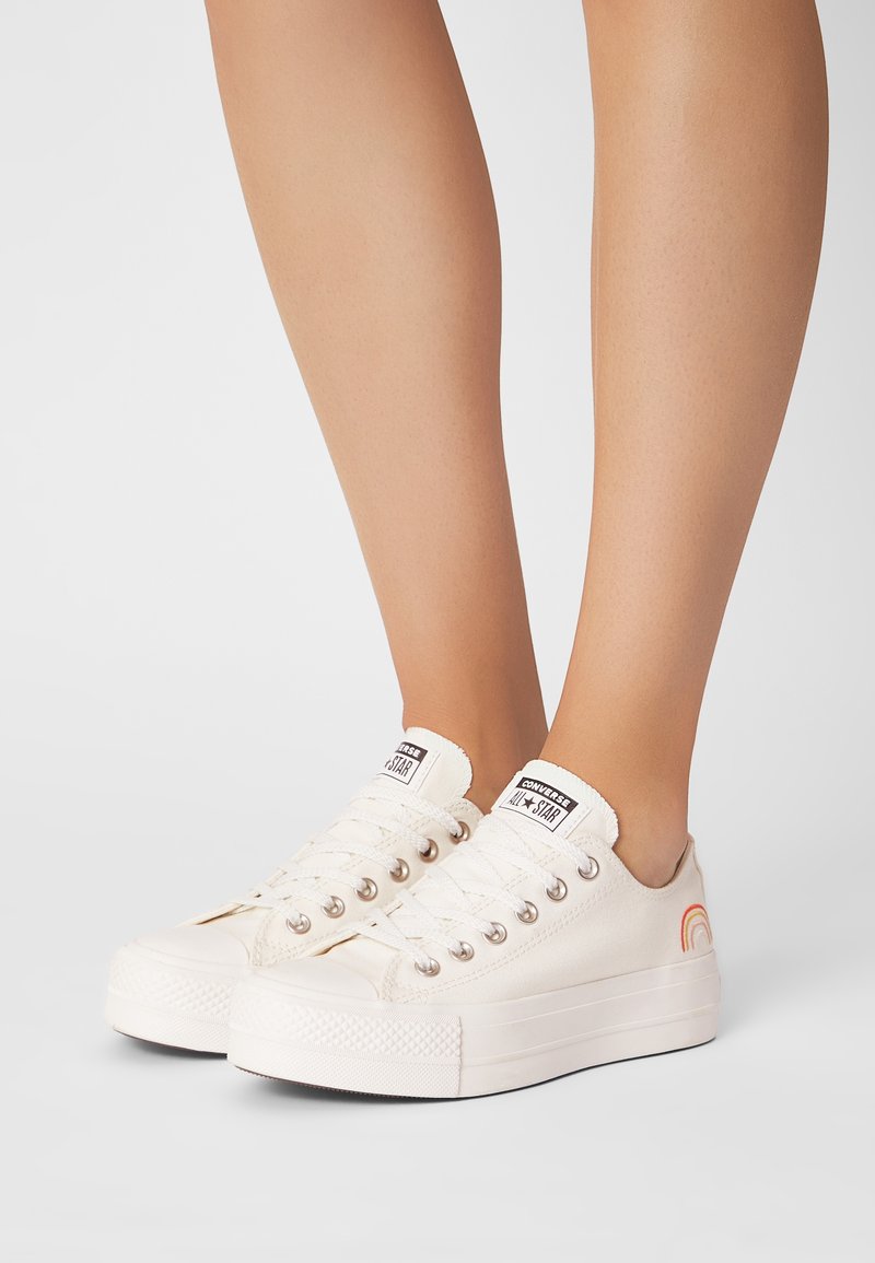 Vita canvas sneakers med lågdesign, silverfärgade öljetter och en regnbågsaccent på sidan. Tjock gummisula med grovt grepp.