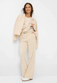 Gilet côtelé beige sur une blouse légère avec détail de volants, associé à un pantalon large crème et des chaussures blanches, sur fond uni.