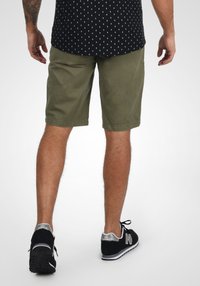 Olivenfarvede shorts lavet af bomuldsblanding, afslappet pasform med forlommer. Bæres med en sort mønstret skjorte og sorte New Balance sneakers.