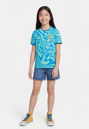 Mädchen lächelt, trägt ein blau-grünes Brasilien-Fußballtrikot, Jeans-Shorts und gelbe High-Top-Sneaker mit blauen Schnürsenkeln, steht vor weißem Hintergrund.