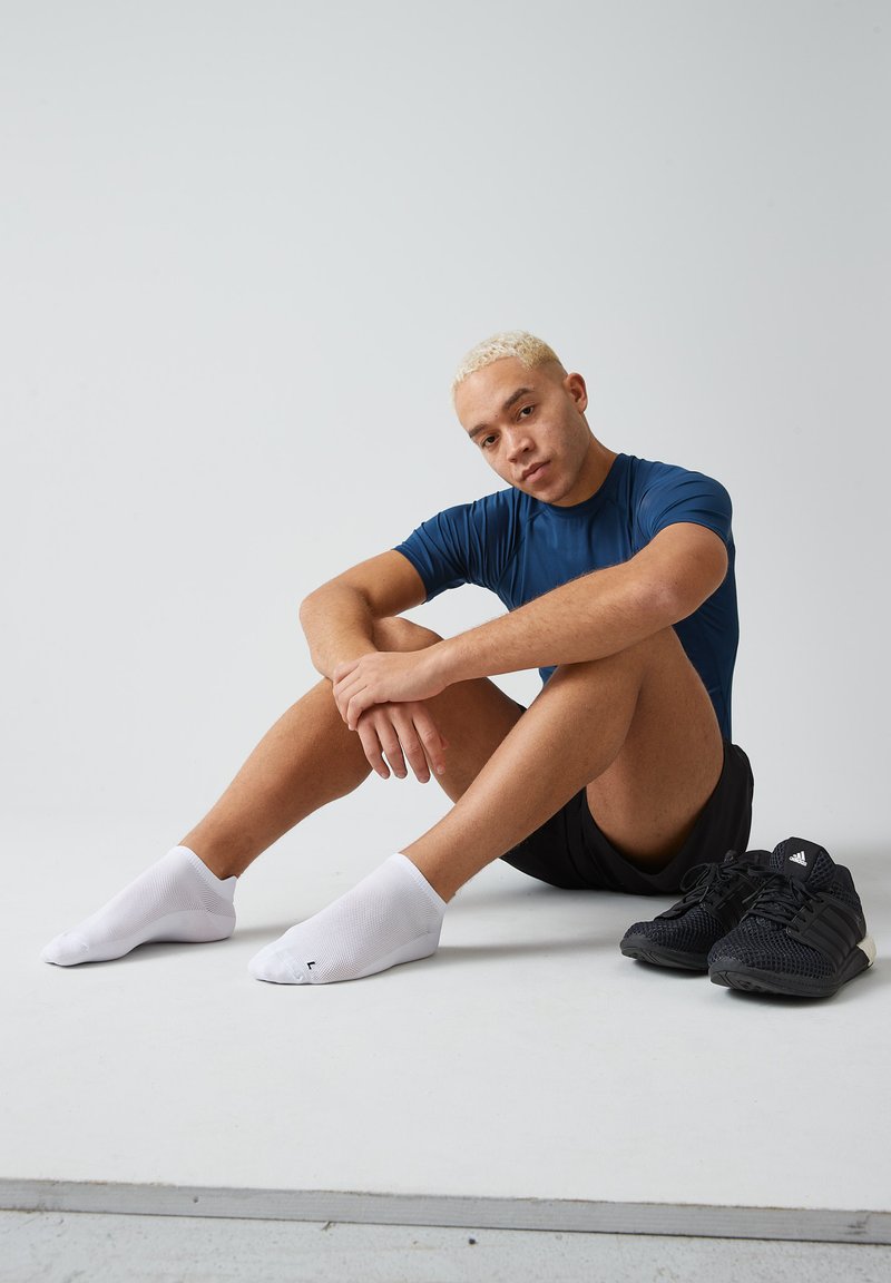 Maglietta atletica blu, pantaloni corti neri, calzini bianchi alla caviglia e scarpe da ginnastica nere con design a rete, posizionati su una superficie grigia liscia.