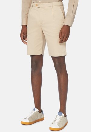 Homme portant un short beige sur mesure, une chemise beige à manches longues et des baskets beige et blanches avec semelles en gomme, debout devant un fond blanc.