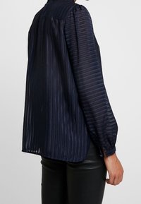 Žena v námořnické modré průhledné blůze s vertikálními pruhy a dlouhými rukávy, kombinovaná s černými koženými kalhotami, zobrazená z boku a zezadu.