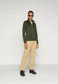 Naine, kandes prille, tumeroheline lukuga jakk, beežid püksid ja mustad kingad, seisab lihtsa heleda tausta ees.