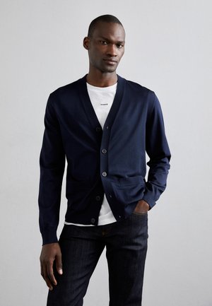 Cárdigan azul marino con escote en V, cierre de botones y bolsillos delanteros, llevado sobre una camiseta blanca. Textura suave y ajuste relajado.