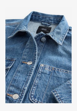 Gai zilo denimu krekls ar klasisko apkakli, priekšējo pogu aizdari, divām krūšu kabatām un sudraba detaļām. Iedobes tekstūra un šūšanas detaļas.