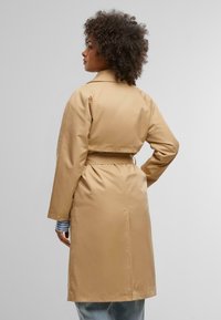 Tan trench coat with a belted waist, long sleeves, and a collar. Features a smooth texture and a mid-length design.