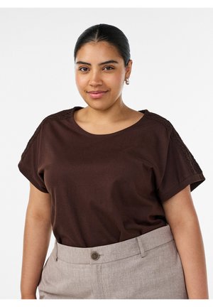 Selvsikker kvinde med mørkt hår bundet tilbage, iført en brun kortærmet top og beige bukser med høj talje, der smiler let mod en hvid baggrund.