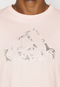 T-shirt en coton rose clair avec un logo Adidas gris texturé présentant trois bandes et un motif de peau de serpent. Design classique à col rond.