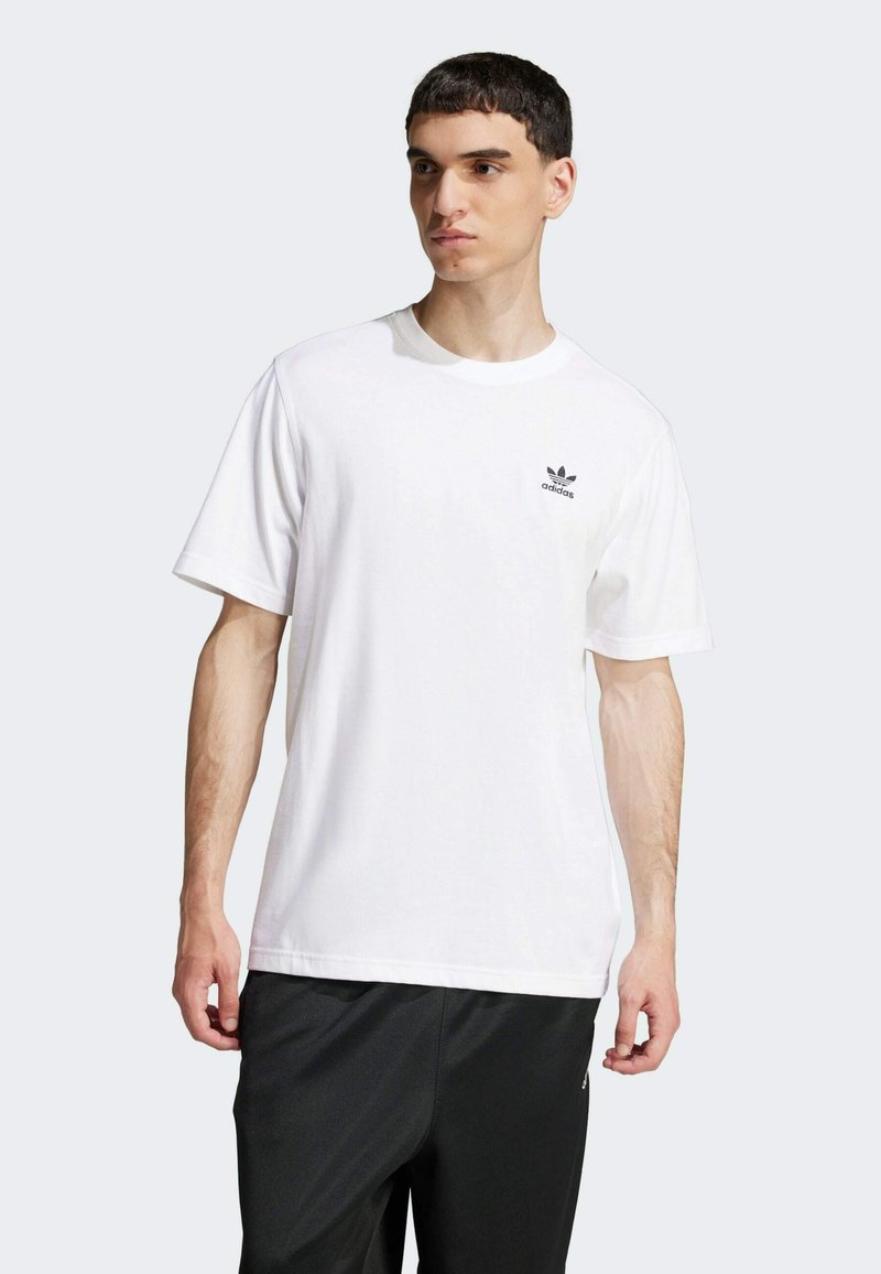Hvid T-shirt i bomuld med korte ærmer, rund hals og et sort Adidas-logo på venstre bryst, kombineret med sorte træningsbukser.