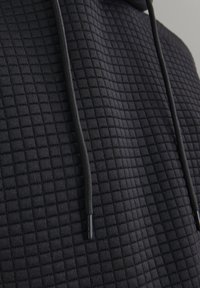 Schwarzer Hoodie mit einem strukturierten, gesteppten Muster, verstellbaren Kordeln und eleganten schwarzen Hardware-Details.