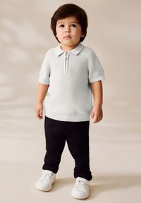 Weißes Poloshirt mit kurzen Ärmeln und Reißverschluss am Kragen, kombiniert mit schwarzer Hose und weißen Sneakers. Weiche Strickstruktur, lässige Passform und gerippte Details.