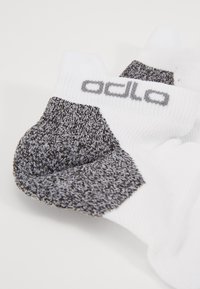 Calcetines deportivos de color blanco y gris con un patrón, con la punta y el talón texturizados en gris, presentando un logo gris en el puño blanco.