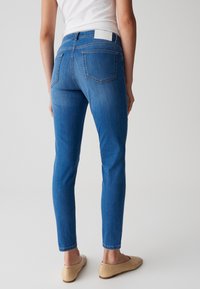 Jean skinny bleu avec une taille mi-haute, présentant deux poches arrière et un délavage clair. Porté avec des ballerines beiges. Texture en denim lisse.