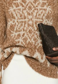 Maglione lavorato a maglia di color marrone chiaro con motivo geometrico crema; il tessuto testurizzato si drappeggia in modo morbido. Clutch nera testurizzata tenuta in mano.