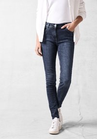 Jean skinny bleu foncé en tissu denim avec une texture lisse, dotée d'une fermeture classique à boutons et à glissière ainsi que de poches avant.