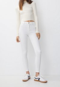 Jean skinny blanc taille haute, haut court à manches longues côtelé couleur crème, et baskets blanches avec accents noirs et semelles en gomme.