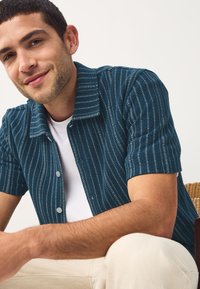 Short-sleeve shirt in dark teal with vertical white stripes, featuring a button-up front and a collared design, made from a textured material.