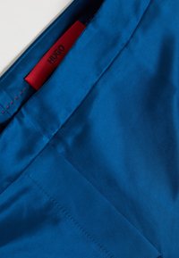 Blue satin fabric featuring a smooth texture, with a red label partially visible. Stitching detail along the seam is also present.