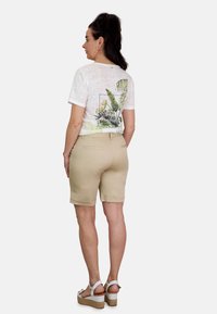 Camiseta blanca texturizada con estampado de hojas verdes en la parte trasera, combinada con pantalones cortos beige hasta la rodilla y sandalias blancas de plataforma.