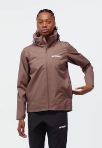 Nainen säätää ruskean Terrex-vetoketjullisen takin hihaa, pukeutuneena mustiin Terrex-housuihin, yksinkertaisen valkoisen taustan edessä.