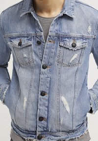 Ljusblå denimjacka med slitna detaljer, knäppning framtill, två bröstfickor och en något fransig fåll.