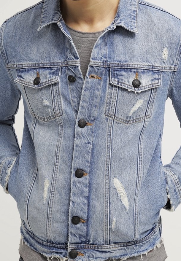 Ljusblå denimjacka med slitna detaljer, knäppning framtill, två bröstfickor och en något fransig fåll.