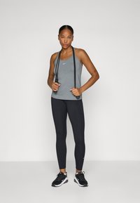 Grå atletisk tank topp med logo, svarte leggings, svarte joggesko med såle accenter, holder et hoppetau, står mot en ensfarget bakgrunn.