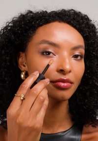 Femme aux cheveux bouclés appliquant de l'anti-cernes sous son œil avec un pinceau de maquillage, portant des boucles d'oreilles créoles dorées et un haut noir.