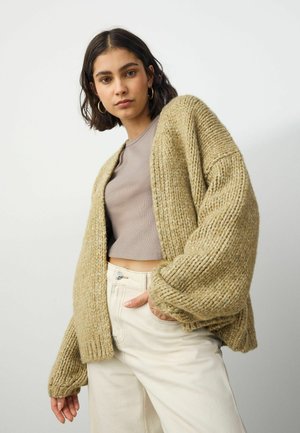 Cardigan en tricot beige avec une texture épaisse, à l'avant ouvert et à manches larges, porté par-dessus un haut gris court et un pantalon crème.