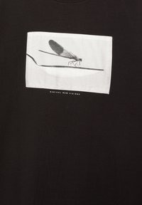 Sort bomulds T-shirt med et hvidt rektangulært grafik af en guldsmed på en ske, med teksten "RADICAL NEW VISIONS" nedenfor.