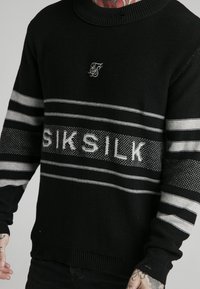 Svart stickad tröja med rund halsringning, grå ränder och "SIKSILK" text i mitten. Ribbad ärm- och nederkant, med en strukturerad finish.